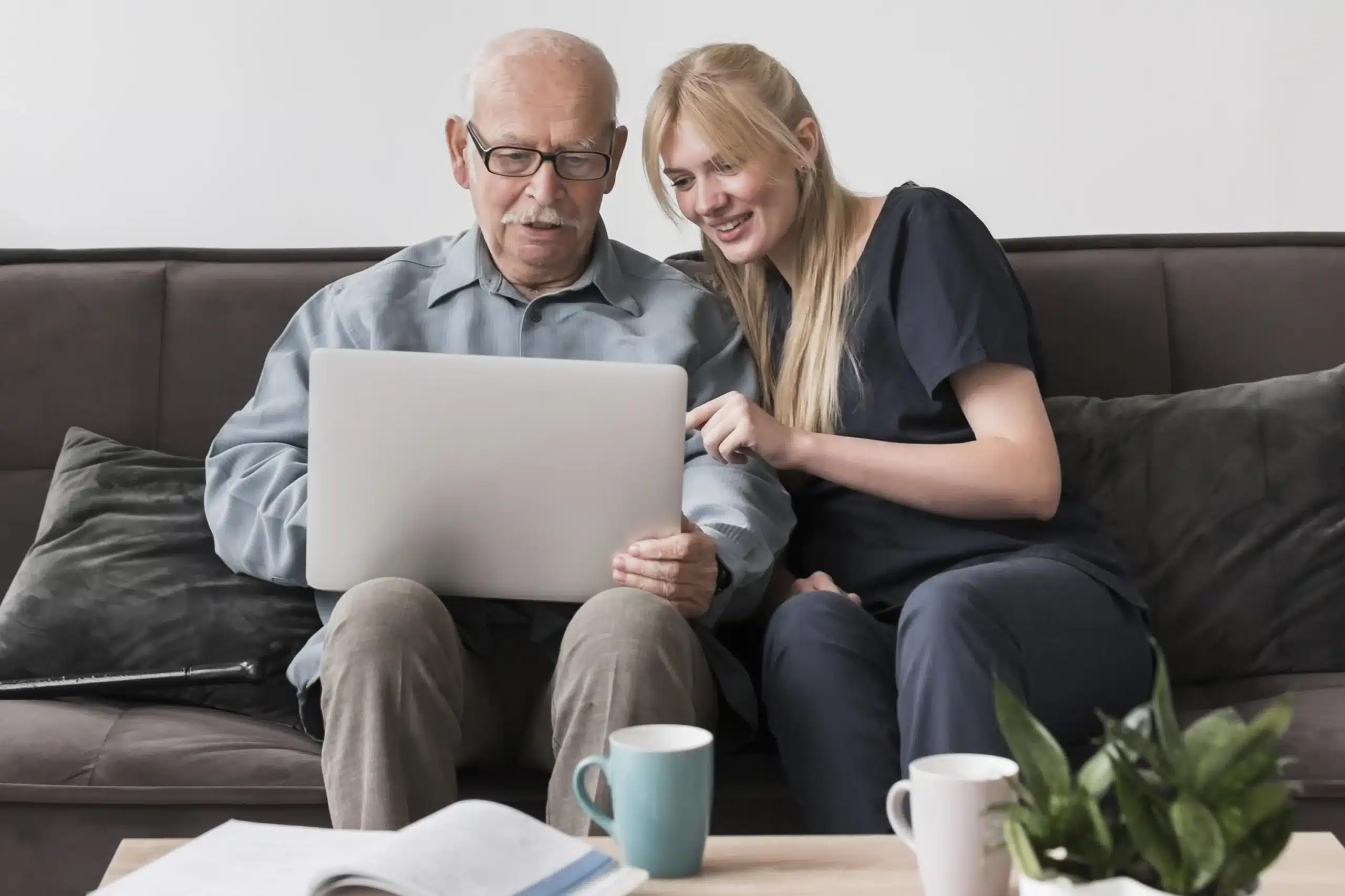 Elderly man receiving digital assistance from a young caregiver, engaging with a laptop in a comfortable home setting
